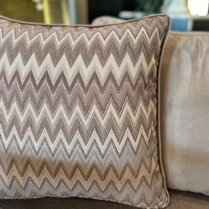May include: A brown and cream chevron patterned throw pillow with a subtle sheen. The pillow is made of a soft, textured fabric.