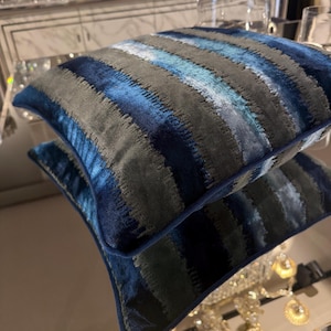 Puede incluir: Dos cojines decorativos con un estampado de rayas en tonos azules y grises. Los cojines tienen una textura suave y aterciopelada y un borde azul oscuro. El diseño presenta rayas horizontales de diferentes anchos.