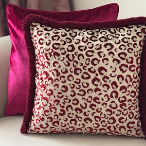 Puede incluir: Dos cojines decorativos. El cojín delantero tiene un fondo crema con un estampado de leopardo rojo y un borde de flecos rojos. El cojín trasero es de color magenta liso. Ambos cojines están hechos de un material suave, similar al terciopelo.