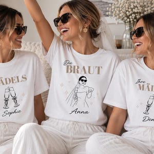 Op de afbeelding: Drie witte t-shirts met grafische prints. Het middelste shirt toont een illustratie van een bruid en de tekst "Die BRAUT" met de naam "Anna". De twee shirts aan de zijkant tonen champagneglazen en de tekst "Ihre MÄDELS" met de namen "Sophie" en "Denise".