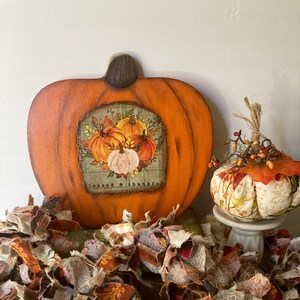 May include: A wooden pumpkin painted orange with a painted image of pumpkins and the text "blessed & thankful". A small fabric pumpkin with a floral pattern sits on a white pedestal. A fabric garland with a patchwork pattern of brown, orange, and white fabric is in the foreground.