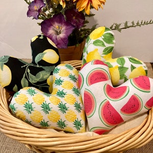 May include: Four heart-shaped fabric pillows in a wicker basket. The pillows are decorated with different fruit patterns: lemons, pineapples, and watermelon.