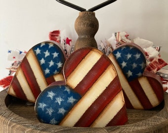 May include: Three wooden heart-shaped ornaments painted with the American flag design. The hearts are displayed on a tiered wooden stand with a black metal handle.