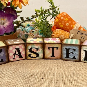 May include: Wooden blocks spelling "EASTER" with distressed edges and various patterns. The blocks are arranged in front of fabric carrots with a "Carrots 5¢" sign and a floral arrangement, creating a festive Easter display.