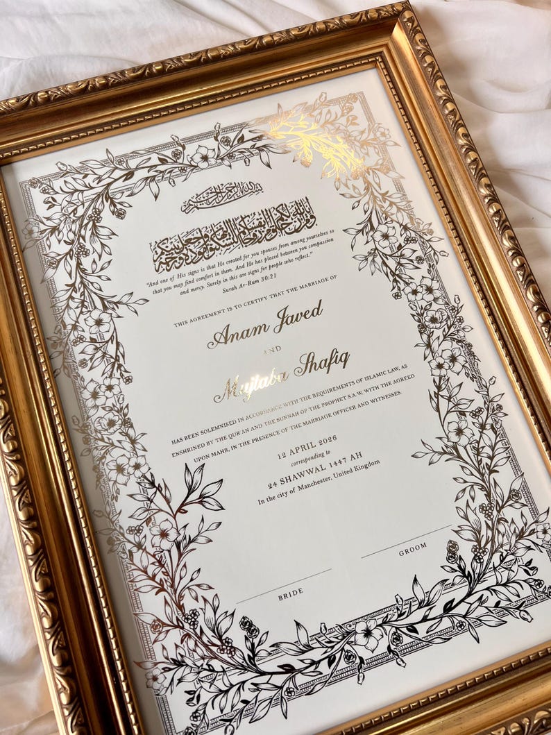 May include: A framed marriage certificate with gold accents. The document features ornate floral border designs and Arabic calligraphy. The names "Anam Javed" and "Mujtaba Shafiq" are prominently displayed. The frame is a gold color.