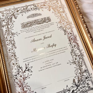 May include: A framed marriage certificate with gold accents and floral designs. The document features elegant calligraphy and the names Anam Javed and Mujtaba Shafiq. The frame is a gold color.