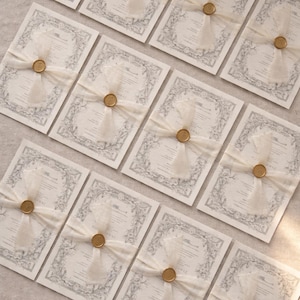 May include: Elegant wedding invitations with a floral border design. Each invitation is tied with a sheer ivory ribbon and sealed with a gold wax seal. The invitations are arranged on a neutral surface.