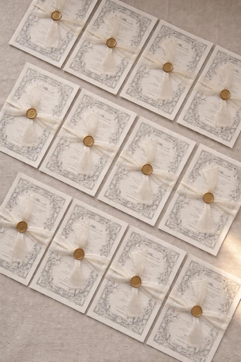 May include: Elegant wedding invitations with a floral border design. Each invitation is tied with a sheer ivory ribbon and sealed with a gold wax seal. The invitations are arranged on a neutral surface.