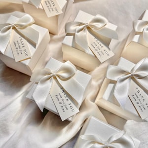 Peut inclure: Une collection de boîtes cadeaux carrées blanc cassé avec une finition texturée. Chaque boîte est nouée avec un ruban de satin couleur crème et comporte une petite étiquette rectangulaire avec le texte : THANK YOU with love from Adam & Asma.