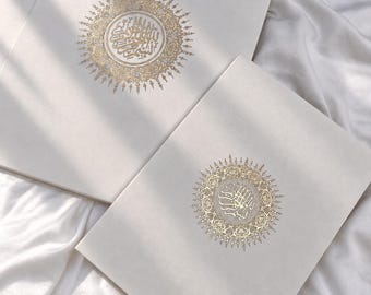 Puede incluir: Dos libros o diarios rectangulares de color blanco roto con un emblema circular de lámina dorada en la portada. El emblema contiene una intrincada caligrafía árabe rodeada por un detallado patrón de rayos de sol en tinta dorada.