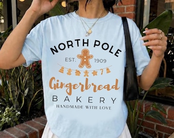 May include: A light blue t-shirt featuring a graphic with the text NORTH POLE EST. 1909 Gingerbread BAKERY HANDMADE WITH LOVE, a gingerbread person icon, and a string of small holiday-themed cookie shapes in brown and black ink.