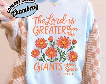 May include: A light blue t-shirt featuring the orange text "The Lord is GREATER than the GIANTS you face" and "1 JOHN 4:4" above and below a graphic of orange flowers with green stems. A Comfort Colors Chambray logo is visible in the corner.