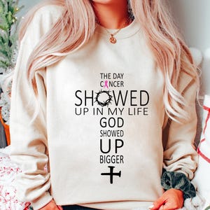 May include: Cream-colored sweatshirt with a cross design. The cross includes the text "THE DAY CANCER SHOWED UP IN MY LIFE GOD SHOWED UP BIGGER" in black, with a pink ribbon and a crown of thorns.
