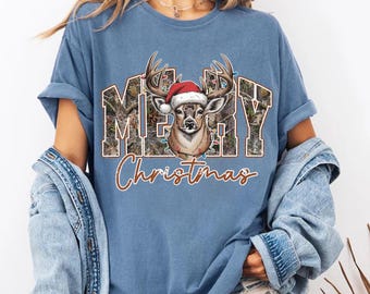 May include: A blue short-sleeve t-shirt featuring a graphic of a deer wearing a red Santa hat. The text MERRY is printed in a block font with a camouflage pattern, and the word Christmas is written below in a brown script font.