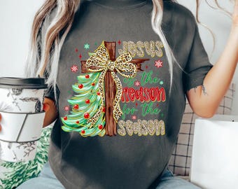 May include: A dark gray t-shirt featuring a graphic of a wooden cross decorated with a leopard print bow and a partial Christmas tree with red ornaments. The text reads "Jesus is the reason for the season" in a mix of leopard print and red fonts.