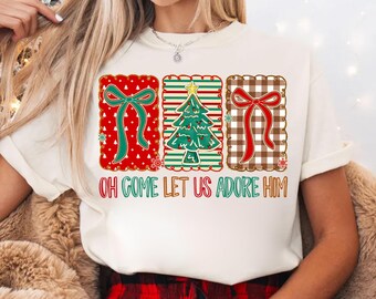 May include: An ivory t-shirt featuring three rectangular panels with scalloped edges. The left panel is red with a green bow, the center is striped with a Christmas tree, and the right is brown gingham with a red bow. Text reads: OH COME LET US ADORE HIM.