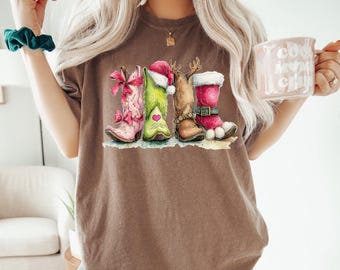 May include: A brown t-shirt featuring a graphic print of four cowboy boots decorated for the holidays. The boots include a pink one with bows, a green one with a Santa hat, a tan one with antlers, and a red one with white fur trim and a buckle.