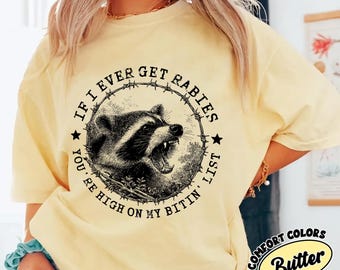 May include: A pale yellow t-shirt featuring a black graphic of a snarling raccoon enclosed within a barbed wire circle. The text reads: IF I EVER GET RABIES YOU'RE HIGH ON MY BITIN' LIST. The shirt has short sleeves and a crew neckline.