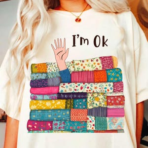 May include: White t-shirt with a colorful illustration of a hand reaching out from a stack of colorful patterned fabric. The text "I'm Ok" is printed on the shirt.