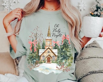 May include: A light green t-shirt featuring a graphic of a church with a steeple, surrounded by pine trees, red bows, and snow. The text "Oh Holy Night" is printed in black script above the church. The design includes festive winter elements.