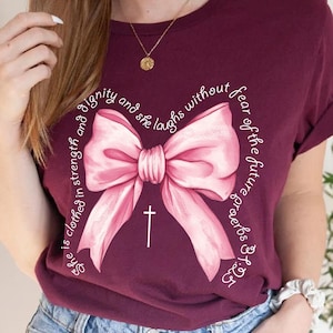May include: A maroon t-shirt with a pink bow and a white cross. The shirt has the text "She is clothed in strength and dignity and she laughs without fear of the future Proverbs 31:25" printed on it.