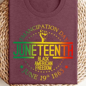 Pode incluir: Camiseta bordô com um gráfico colorido que celebra o Juneteenth. O design inclui a palavra "Juneteenth" em um gradiente de verde, amarelo e vermelho, com o texto "Black American Freedom" e "June 19th 1865". Um gráfico de punho erguido também está incluído.
