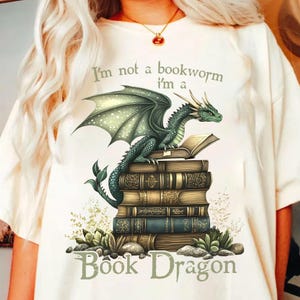 May include: A white t-shirt with a green dragon sitting on a stack of books. The dragon is holding a book in its claws. The text on the shirt reads "I'm not a bookworm I'm a Book Dragon".