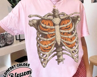 May include: A light pink t-shirt featuring a graphic illustration of a human rib cage containing several orange jack-o'-lantern pumpkins. The design is centered on the chest area of the shirt, which has a crew neckline and short sleeves.