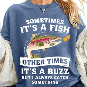 May include: A blue t-shirt with a white graphic of a fish with a beer can in its mouth. The text on the shirt reads "Sometimes it's a fish, other times it's a buzz, but I always catch something."