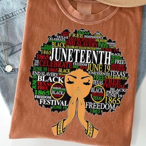 May include: A brown t-shirt featuring a graphic design celebrating Juneteenth. The design includes the text "Juneteenth" in large letters, along with other words and phrases such as "Freedom", "Black", and "Texas". The graphic also features an illustration of a woman with her hands clasped in prayer.