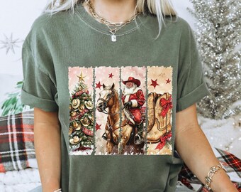 May include: An olive green crew neck t-shirt featuring a graphic print divided into three vertical panels. The panels display a decorated Christmas tree, Santa Claus riding a brown horse, and a pair of tan cowboy boots with red bows.
