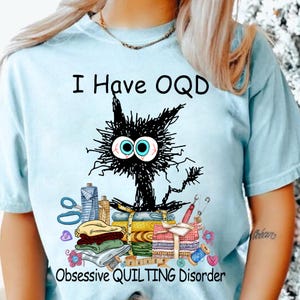 Puede incluir: Una camiseta azul claro con una ilustración de un gato negro y el texto "I Have OQD Obsessive Quilting Disorder".