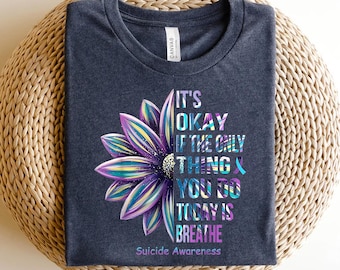 May include: A dark gray t-shirt with a purple and blue flower graphic. The text on the shirt reads "It's okay if the only thing you do today is breathe. Suicide Awareness."