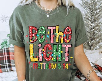 May include: An olive green short-sleeve t-shirt featuring the text BE THE LIGHT MATTHEW 5:14 in large, colorful, polka-dotted block letters. The design is accented with a string of multi-colored Christmas lights wrapped around the lettering.