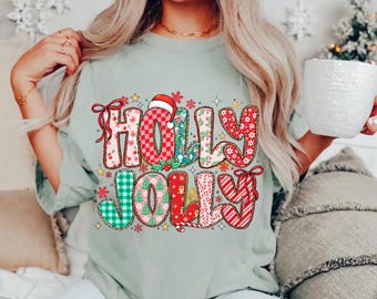 May include: A light sage green t-shirt featuring a large, colorful graphic print with the text HOLLY JOLLY. The letters are decorated with various holiday patterns including red and white checks, green plaid, candy cane stripes, and small Christmas trees.