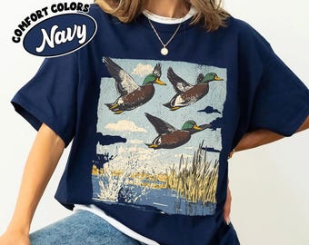 May include: A navy blue t-shirt featuring a graphic print of three mallard ducks in flight over a marshy landscape with tall reeds and water. The text COMFORT COLORS Navy is printed in the upper left corner of the shirt in a circular design.