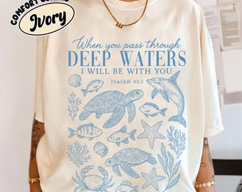 May include: An ivory t-shirt featuring blue line art of sea creatures including turtles, a dolphin, a crab, fish, starfish, and coral. The text reads: When you pass through DEEP WATERS I WILL BE WITH YOU ISAIAH 43:2. A gold chain necklace is worn.