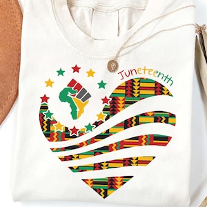Peut inclure: T-shirt crème avec un motif en forme de cœur avec un imprimé africain coloré. Le motif comprend un poing levé, une carte de l'Afrique et des étoiles. Le mot "Juneteenth" est écrit en vert, rouge et jaune. Un collier en or est drapé sur le t-shirt.