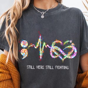 May include: Dark gray t-shirt with a colorful graphic design featuring a semicolon, heartbeat, and infinity symbol. The words "STILL HERE STILL FIGHTING" are printed below the graphic. The design incorporates rainbow colors.