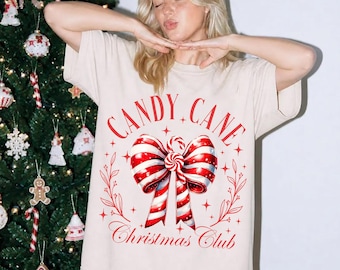 May include: An ivory t-shirt featuring a red graphic print of a large candy cane striped bow in the center. The text reads Candy Cane Christmas Club in a red arched font. The design includes small star accents and thin leafy branch illustrations.