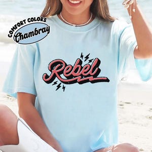 May include: Light blue t-shirt with the word "Rebel" in a retro font, outlined in black and pink, with lightning bolt accents. The shirt also features the text "Comfort Colors Chambray" in a circular design.