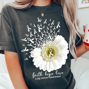 Puede incluir: Camiseta gris oscuro con una margarita blanca y cintas blancas. El texto "faith hope love" y "Lung cancer awareness" están impresos debajo. Un diseño para concienciar sobre el cáncer de pulmón.