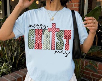 May include: A light blue short-sleeve t-shirt featuring the text "merry CHRIST mas" in a red and green checkered pattern. The letter "T" in "CHRIST" is stylized as a cross. The shirt has a crew neckline and a relaxed fit.