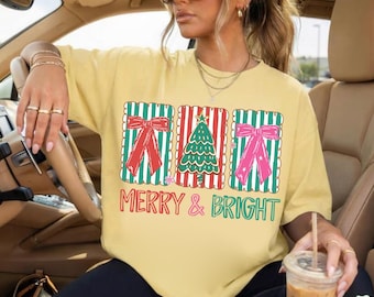May include: A pale yellow t-shirt featuring a graphic of three rectangular panels with red and green stripes. The outer panels show red and pink bows, while the center panel displays a green Christmas tree. The text MERRY & BRIGHT is printed below.