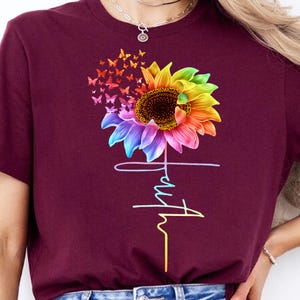 May include: A maroon t-shirt with a rainbow sunflower and butterflies. The word "truth" is written vertically below the sunflower.