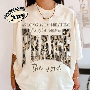 May include: An ivory t-shirt featuring the text "AS LONG AS I'M BREATHING I've got a reason to PRAISE the Lord". The word "PRAISE" is printed in large, block letters with a brown and black leopard print pattern, while the other text is in black.