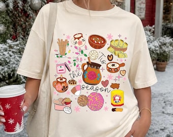 May include: An ivory short-sleeve t-shirt featuring a colorful graphic print of various Mexican holiday foods and drinks, including a clay pitcher, tamales, and sweet bread, with the text 'Tis the season' written in black cursive script.