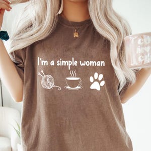 Puede incluir: Una camiseta marrón con el texto "Soy una mujer sencilla" y tres iconos: una bola de hilo con agujas de tejer, una taza de café y una huella de pata.