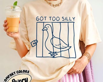 May include: An ivory short-sleeved t-shirt featuring a dark blue line art graphic of a duck inside a cage with a padlock. The text above the cage reads GOT TOO SILLY. The shirt has a crew neckline and is worn with a gold chain necklace.