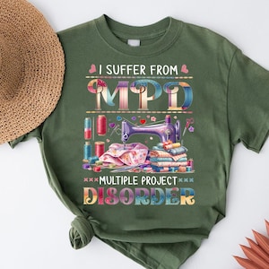 Może przedstawiać: Zielona koszulka z napisem "I suffer from MPD Multiple Project Disorder". Projekt zawiera maszynę do szycia, szpulki nici, tkaninę i nożyczki. Słomkowy kapelusz jest widoczny w lewym górnym rogu.